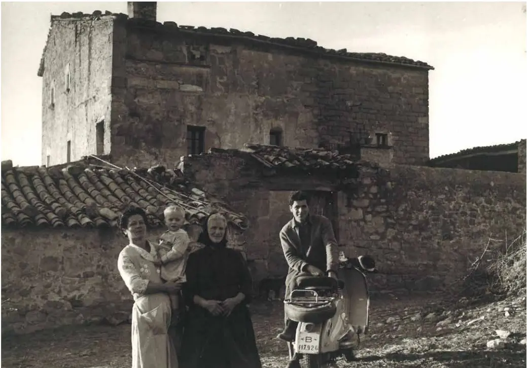Fotografia antiga: una família davant d'una casa de pedra, amb una moto Vespa